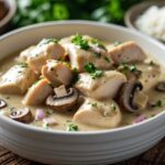 Prato de strogonoff de frango cremoso servido em uma tigela branca sobre uma mesa de madeira, acompanhado de arroz branco e ervas frescas ao fundo.