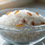 Tigela de vidro transparente com arroz branco e larvas visíveis entre os grãos sobre uma bancada de cozinha.
