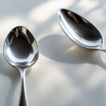 Duas colheres de metal, uma de chá menor e uma de café maior, lado a lado sobre fundo neutro.