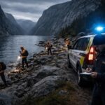 Equipe de busca e resgate em roupas de alta visibilidade procurando na costa rochosa de um fiorde na Noruega sob um céu nublado.